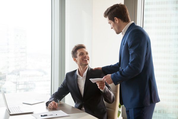 A standing person hands an envelope to a smiling person seated at a desk.